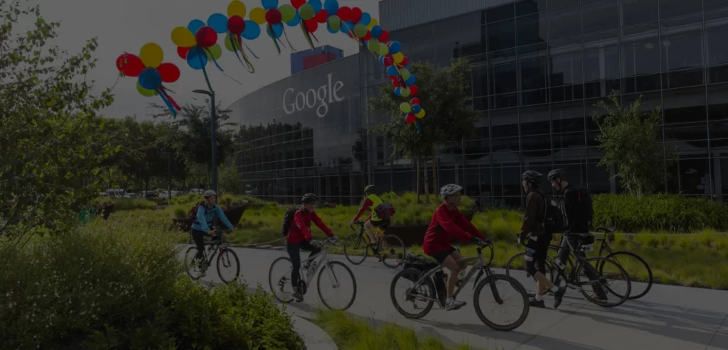 Image of the Office of Google, containing some people riding bikes and holding to colorful balloons